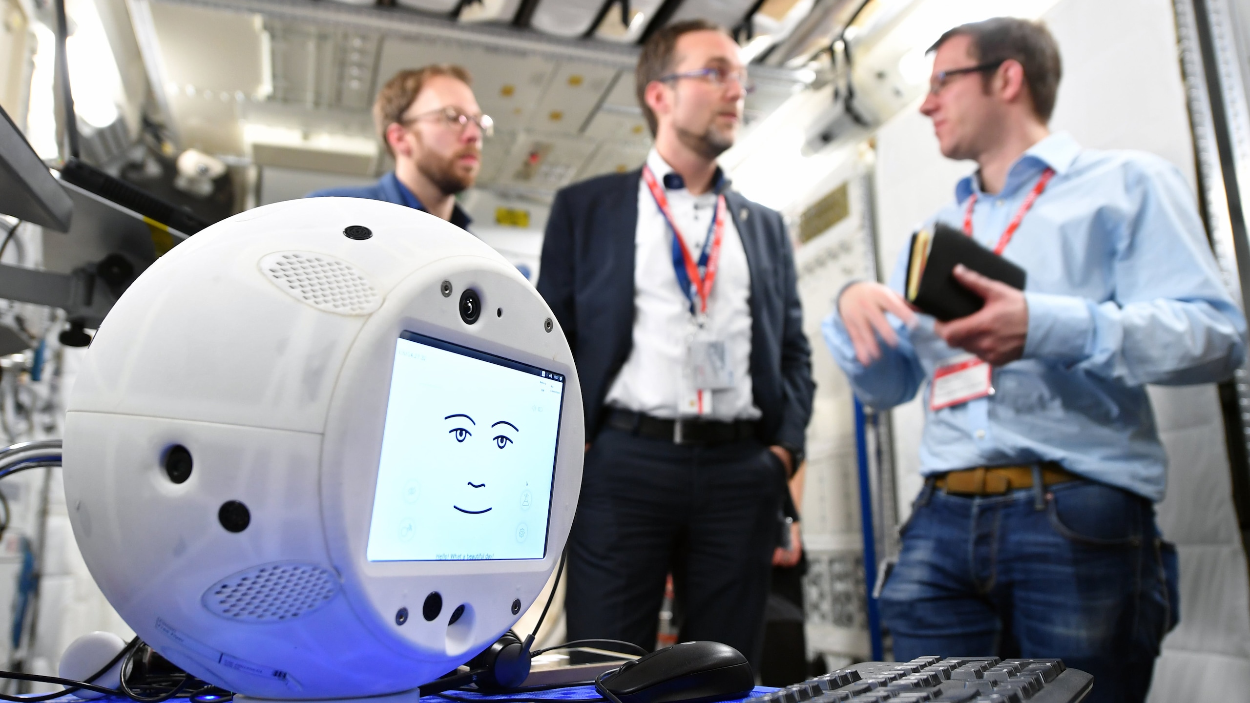 CIMON, the intelligent astronaut assistant, sits in the foreground with three men talking in the background.